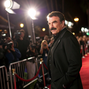 Tom Selleck, mustached actor, in black suit. Poses against a bustling crowd backdrop. Bright lights and camera flashes