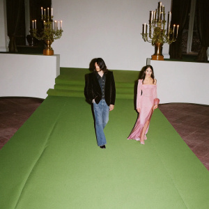 Gilby Clarke and Daniella Clarke attending a high-end fashion gala. The venue features a glamorous, dimly lit stage with chandeliers. They are both dressed in contemporary outfits, with Gilby in a sleek black blazer and jeans.