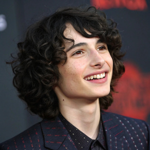 Close-up portrait of Finn Wolfhard, curly dark hair, warm smile, wearing a dark textured blazer with subtle pattern. Dark blurred background with hint of red. Soft, even lighting.