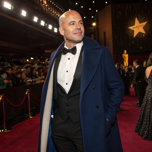 Billy Zane, fair-skinned and bald, in formal wear. Wearing a black vest and crisp white shirt. Dark blue coat draped over his shoulders. At a glamorous awards ceremony. The dimly lit room filled with celebrities