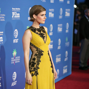 Kate Mara standing confidently in a glamorous red carpet event. She wears a stunning yellow dress with black lace overlay. Focused expression, standing against a vibrant blue background with film festival logos.