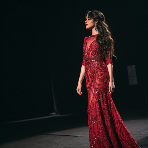 Camila Cabello, standing solo in a spotlight. Wearing a stunning red sequined dress with intricate designs. Dark, minimalist background.