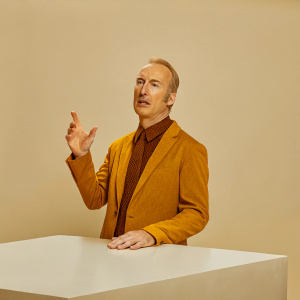Bob Odenkirk, wearing a navy suit with a textured tie, speaks at a tech conference. Minimalist white background