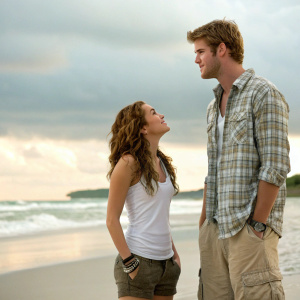 Miley Cyrus and Liam Hemsworth, standing on a beach in The Last Song. Miley in a white tank top with long wavy brown hair, Liam in a plaid shirt and cargo shorts. Neat tousled hairstyle.