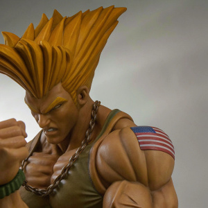 Muscular figurine of Guile from Street Fighter IV. Intense fighting stance, showcasing detailed spiked hairstyle and American flag on arm. Set against a minimalist backdrop.