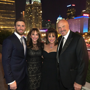 Jay McGraw, Erica Dahm, Robin McGraw, Dr. Phil McGraw at a gala event. They are dressed in formal attire. Dark blazers and dresses. The background is a vibrant cityscape at night.