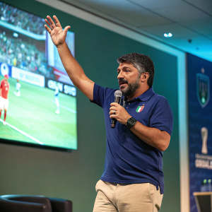 Gennaro Gattuso, an Italian football coach, during a press conference. Wearing a blue polo shirt and khaki pants. Speaking confidently with raised hand. Background includes a large screen showing highlights of a soccer match.