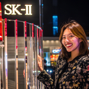 Sooyoung, member of Girls' Generation, smiling in a photoshoot. Wearing a black floral patterned dress. Dark, sleek background with large SK-II logo