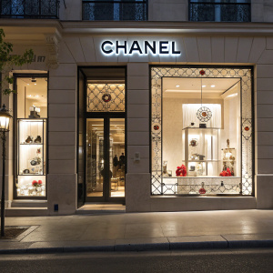 A stunning view of the Chanel boutique at night. Ornate window displays filled with exquisite accessories, illuminated by soft lights. The iconic interlocking C logo stands out prominently above the door.