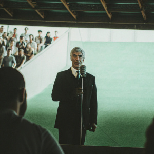 Gian Piero Gasperini, in a formal suit, giving a press conference at the stadium. Background filled with cheering fans and reporters.