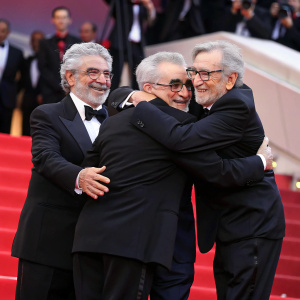 Martin Scorsese, draped in a black tuxedo, is enveloped in a celebratory hug by Francis Ford Coppola, George Lucas, and Steven Spielberg. The quartet stands on a vibrant red carpeted stage, adorned with accolades and flanked by cheering crowds. The atmosphere exudes joy and recognition.
