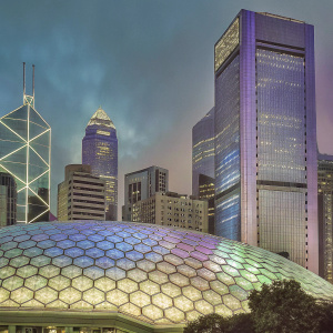 Evening shot of Hong Kong's skyline. Towering buildings and modern structures. Dome-shaped building with hexagonal patterns. Vibrant blue, green, and purple hues from neon lights. Traditional architecture contrasts with modern glass facades.