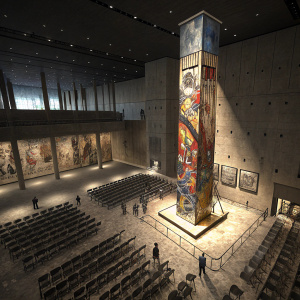 A high-angle shot of the Foundation Hall at the 9/11 Memorial Museum. Rows of chairs fill the space under dim lighting. A tall, colorful column is the focal point, decorated with graffiti-like designs.