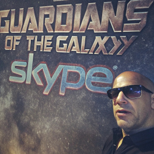 Vin Diesel, wearing a black polo shirt and sunglasses, stands against a colorful backdrop featuring 'Guardians of the Galaxy' and Skype logos. Close-up shot.