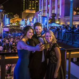 Samantha Mollen, Matt Dallas, and Jenny Mollen at a charity gala. They pose in front of a bustling crowd and vibrant city lights.