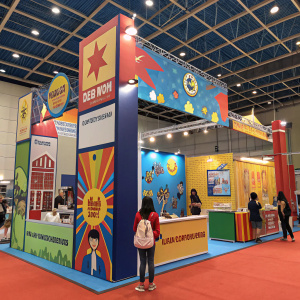 EduHero's vibrant booth at the education expo. Superhero-themed banners, giveaways, and promotional materials attract attendees. Bright, energetic setting.