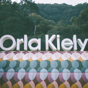 The Orla Kiely logo in sleek white font, placed on a vibrant wallpaper backdrop featuring geometric shapes in various pastel shades.