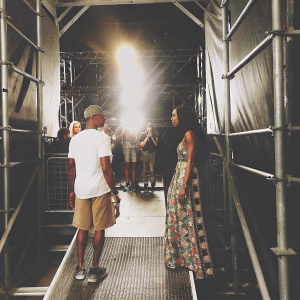Pharrell Williams and Naomi Campbell backstage at a music festival. Bright lights and fans visible in the background. Pharrell in white t-shirt and khaki shorts. Naomi in vibrant patterned dress.