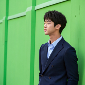 Ha Seok-Jin, an Asian youth with dark hair and a serene expression, dressed in a dark navy suit with a light blue shirt. Standing against a vivid green backdrop with no visible details, looking slightly to the side.