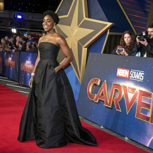 Lashana Lynch, a black actress with curly hair, in a black, strapless gown with a long train. Standing on the red carpet at the Captain Marvel premiere. Behind her, the Marvel Studios logo and cosmic designs. A real-life event with a vibrant atmosphere.