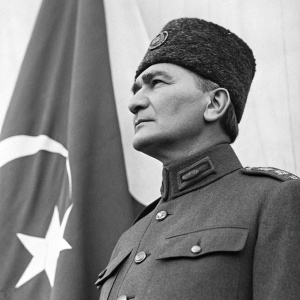 Black and white photo of Mustafa Kemal Atatürk. He wears a military uniform, standing in front of a flag.