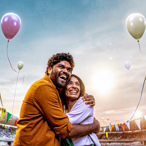 Mark Philippoussis and Amanda Salinas, both with smiles, embrace at a sports award show. The background is a vibrant stadium scene with crowd and banners.