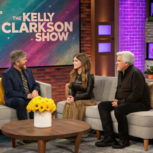 Anna Kendrick, Cam, and Jay Leno are seated on a gray couch. The set features a vibrant backdrop and a brick wall. A table with a yellow flower vase is in the foreground. Studio setting of The Kelly Clarkson Show