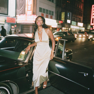 Kimora Lee Simmons, confidently posing at a fashion gala. Wearing an elegant white dress adorned with gold accessories. The backdrop features a bustling street with neon lights.