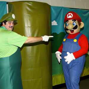 Cartoony depiction of Mario in cheerful pose, pointing at Goomba, set against a bright yellow backdrop.