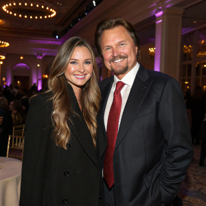 Vanessa Lee Evigan and Greg Evigan at a glamorous event. They both wear smiles. Vanessa in black coat, long hair. Greg in dark suit and red tie.