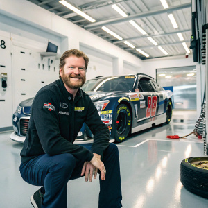 Dale Earnhardt Jr., bearded and smiling, poses with his racing car in a sleek modern garage. High-tech equipment and tools in the background.