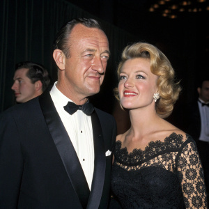 Formal event snapshot. David Niven Jr. and Barbara Niven in elegant attire. David in black tuxedo, Barbara in black lace gown. Dark, sophisticated backdrop
