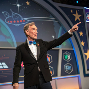 Bill Nye, in a formal outfit, appears on a virtual stage set for a science fiction convention. Wearing a black suit and blue tie, he gestures enthusiastically. The backdrop features the iconic Enterprise and starship insignias. Close-up shot