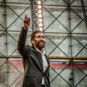 Jake Gyllenhaal, with a full beard, dressed in a dark suit and white shirt, standing on a brightly lit stage. The backdrop features a mix of vibrant colors and geometric shapes.