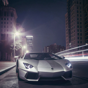 A shiny silver Lamborghini Aventador on a bustling city street at night. Surrounded by tall buildings and bright lights.