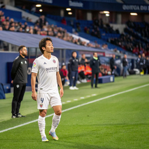 Shinji Okazaki, soccer player for SD Huesca, standing on the vibrant green pitch during a La Liga match at El Sadar Stadium. Wearing his team's white uniform adorned with sponsors' logos. Coaches and staff visible in the background. Spectators in the stadium seating area.