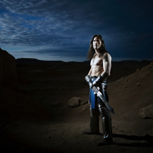 Conan the Barbarian, shirtless with long dark hair, wearing heavy metal armor pieces. Standing amidst a desert wasteland, his expression fierce and determined.