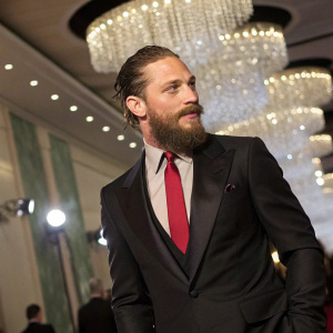 Tom Hardy, known for his rugged looks and thick beard, at a gala event. Wearing a sleek black suit and a deep red tie. Shimmering chandeliers in the background.