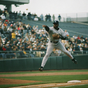 Didi Gregorius, an African American baseball player, in mid-swing. New York Yankees jersey and cap, mid-air with glove catching a ball. Crowd blurred in background, dynamic action shot.