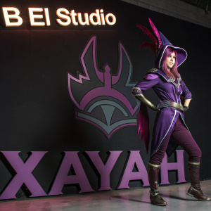 Xayah from League of Legends, in purple attire with a distinctive hat, posing in a shadowy studio. The backdrop features the 'B El Studio' logo and her name in bold text.