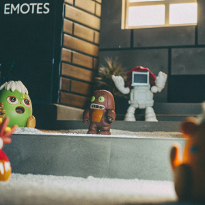 A vibrant, cartoon-style depiction of League of Legends emotes. Features a variety of characters with distinct designs, including a fiery green creature, a brown and gray figure, and a futuristic robot. Set against a stylized grassy backdrop labeled 'EMOTES'