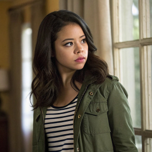 Cierra Ramirez, known for her role as Callie in 'The Fosters', posing seriously. Dark hair, green jacket, striped shirt. Indoor setting with blurred background, window with curtain visible.