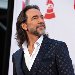 Marco Antonio Solís at the Latin Grammy Awards. Wearing a dark suit with a white shirt underneath. Behind a simple white backdrop.