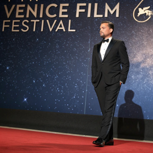 Leonardo DiCaprio at Venice Film Festival premiere. Wearing a black tuxedo. Dark red carpet. Starry night backdrop with festival logos.