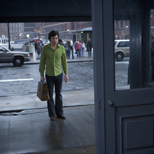 Adam Brody in a vibrant green shirt and dark jeans, carrying a brown bag, standing confidently outside a bustling cityscape.