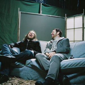 Axl Rose and Jimmy Kimmel sharing a joke on a talk show. Behind the scenes, green screen backdrop, casual attire