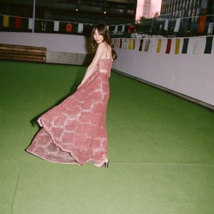 Olga Kurylenko, draped in a flowing red dress with intricate lace patterns, poses gracefully against the backdrop of a bustling city street festival. The vibrant scene includes colorful banners and lively music.
