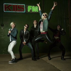 Ross Lynch, Ellington Ratliffe, Riker Lynch, Rydel Lynch, Rocky Lynch, and R5 performing at a KIIS FM event. The stage is set against a bright neon backdrop with bold logos and text. They wear casual yet stylish outfits, including denim and leather jackets.