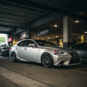 Silver Lexus IS350 F Sport in a dimly lit garage. Various cars and dealership signage visible.