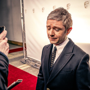 Martin Freeman, wearing a dark suit and tie, standing on a red carpet at the BAFTA TV Awards. Against a plain white backdrop with event logos. Sole focus on the actor.
